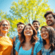 A group of happy people outdoors on a sunny day, smiling under clear blue skies with green trees, radiating positive energy and joy.