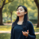 Young adult woman practicing deep breathing in a peaceful park with soft natural light and lush greenery, symbolizing relaxation and anxiety relief.